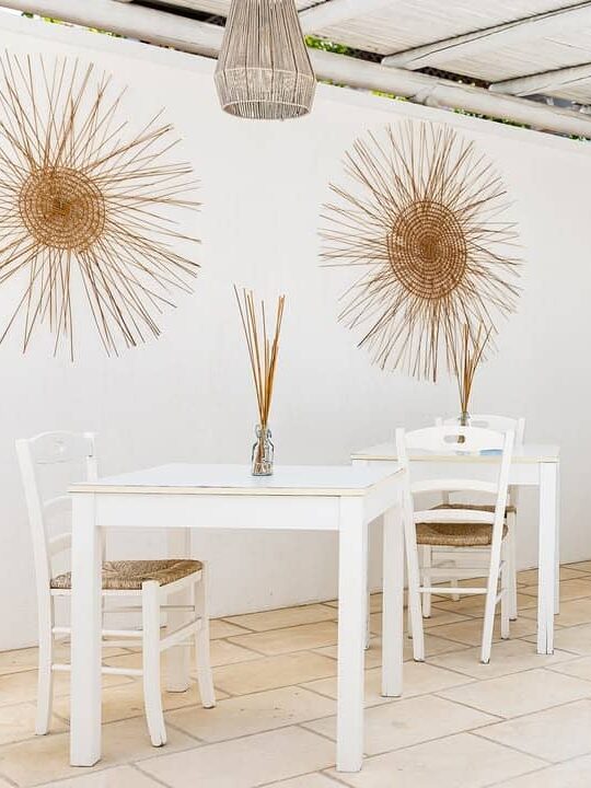 White minimalist cafe with woven wall art, white tables and chairs, and natural light from a covered patio&mdash;evoking the relaxed elegance found at the best beach resorts in Italy for families.