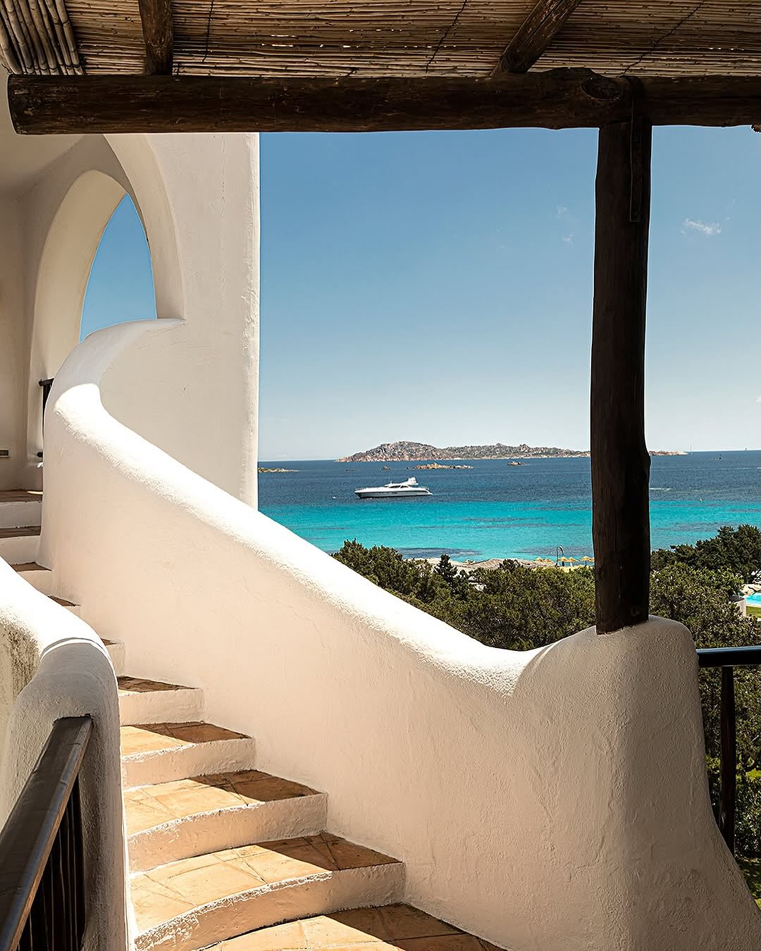 White curved staircase with terracotta tiles overlooks turquoise sea and a yacht under a clear blue sky, capturing the serene charm found at the best beach resorts in Italy for families.