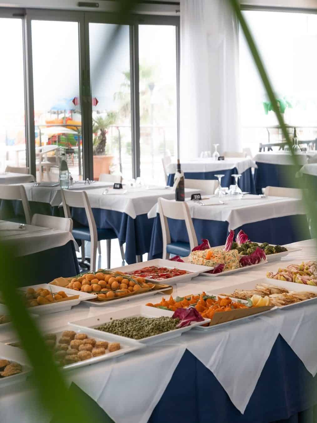 Buffet table with assorted food dishes in a bright restaurant, reminiscent of the dining experiences at the best beach resorts in Italy for families, with neatly set tables and large windows.