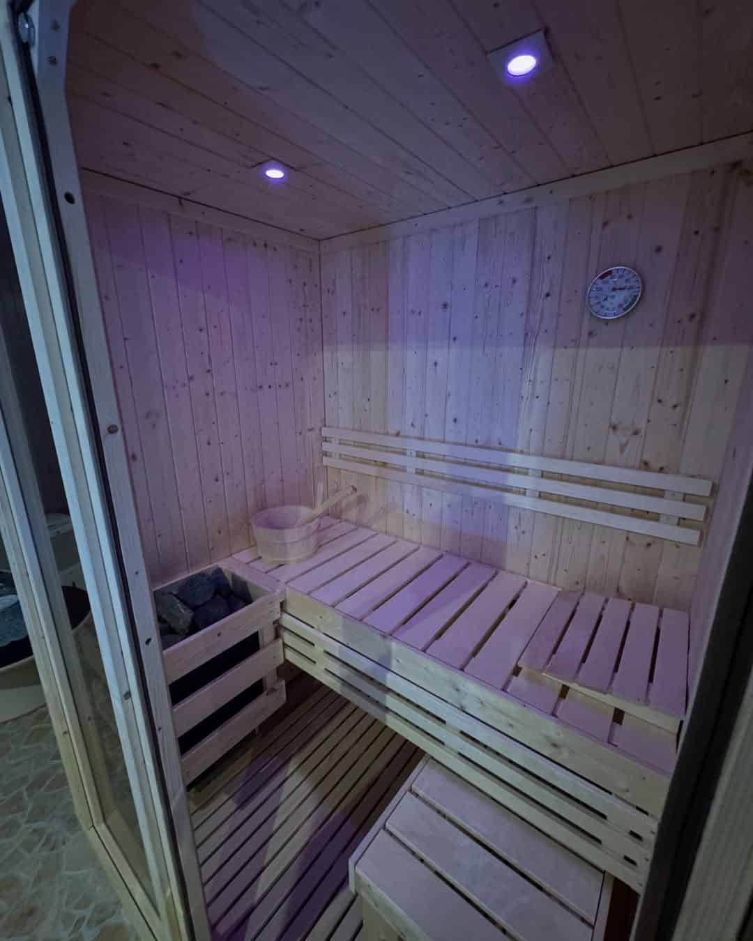 Small wooden sauna room with bench seating, a bucket, stones, and a wall-mounted thermometer&mdash;an inviting spot reminiscent of relaxing amenities found at the best beach resorts in Italy for families.