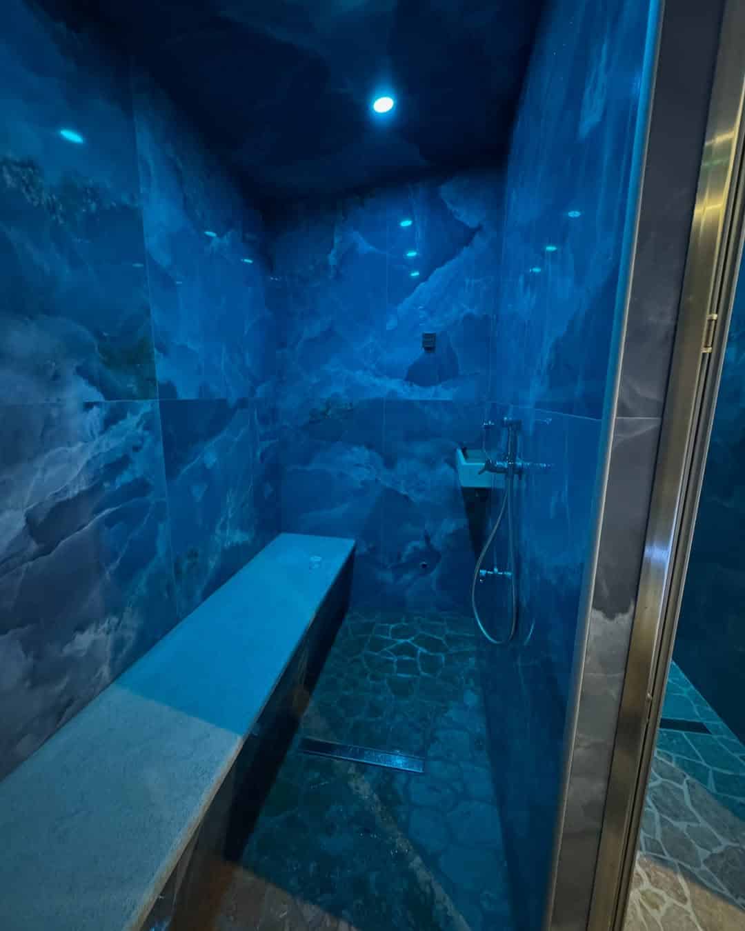 Blue-tiled steam room with a stone bench, rainfall shower, and a ceiling-mounted light&mdash;offering relaxation reminiscent of the best beach resorts in Italy for families.