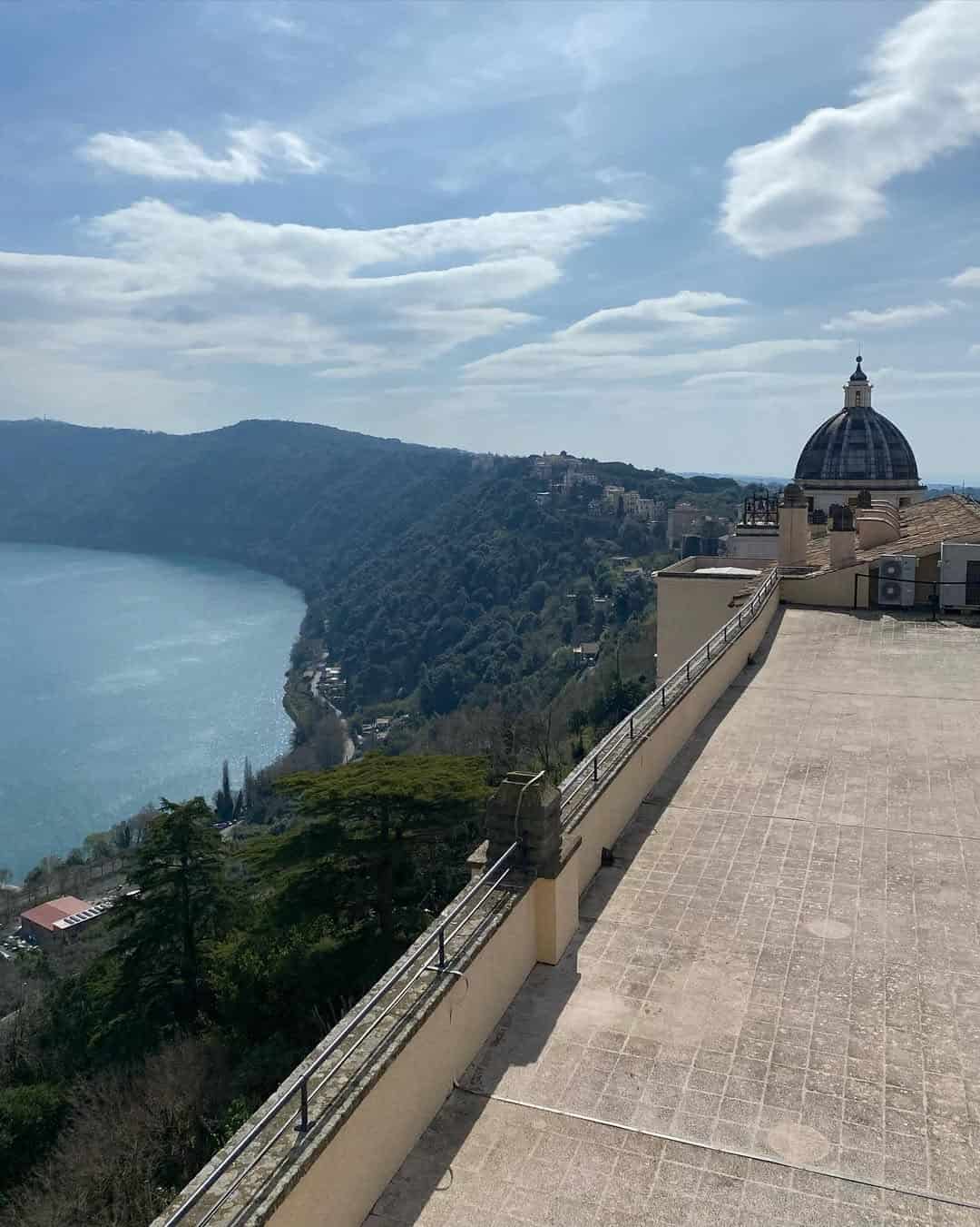 A domed building overlooks a lake and forested hills under a partly cloudy sky, capturing the serene beauty you'll experience during 4 days in Castel Gandolfo.