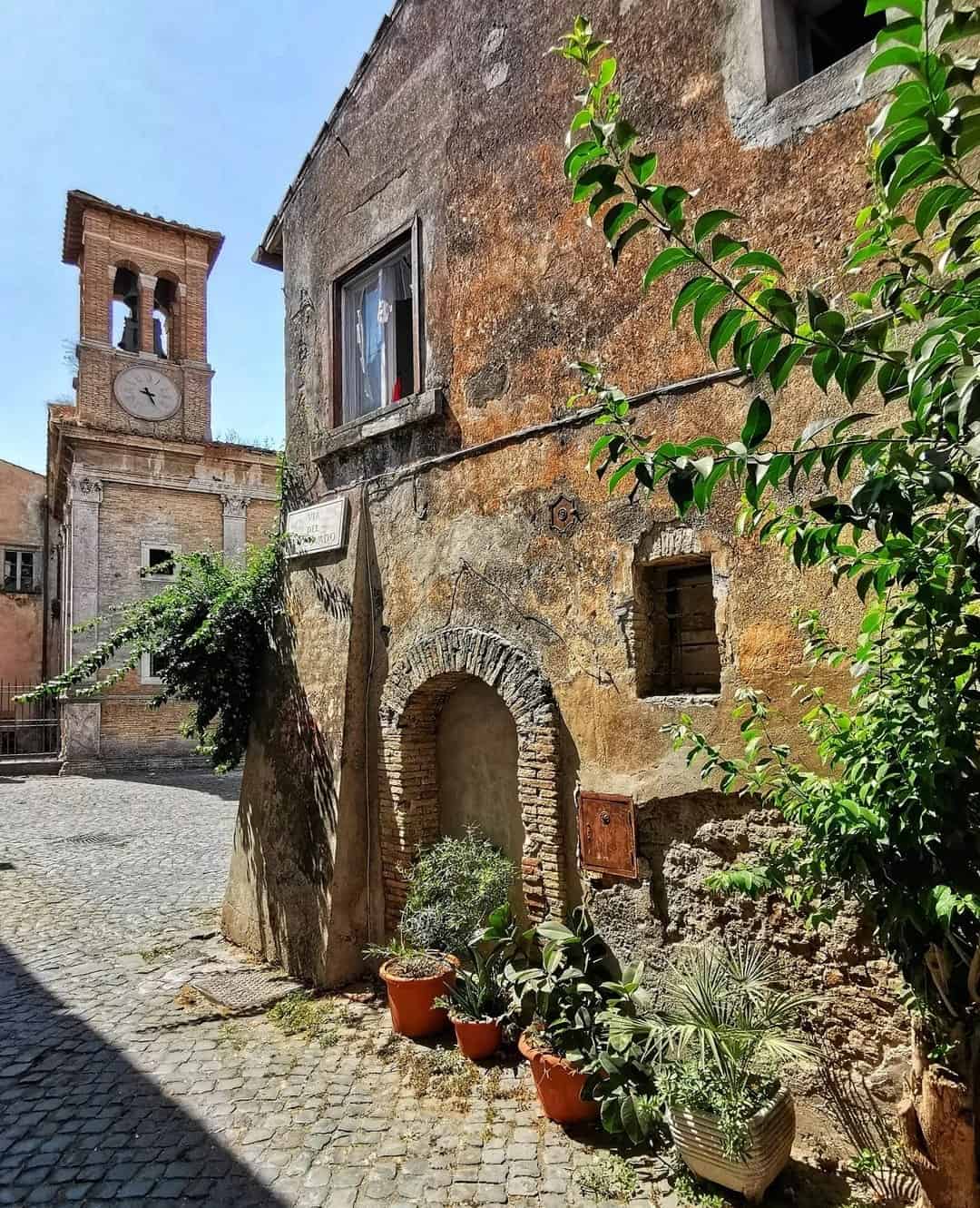 A sunlit cobblestone street with old stone buildings, potted plants, and a clock tower in the background&mdash;scenes like this await on the best day trips from Rome for families.