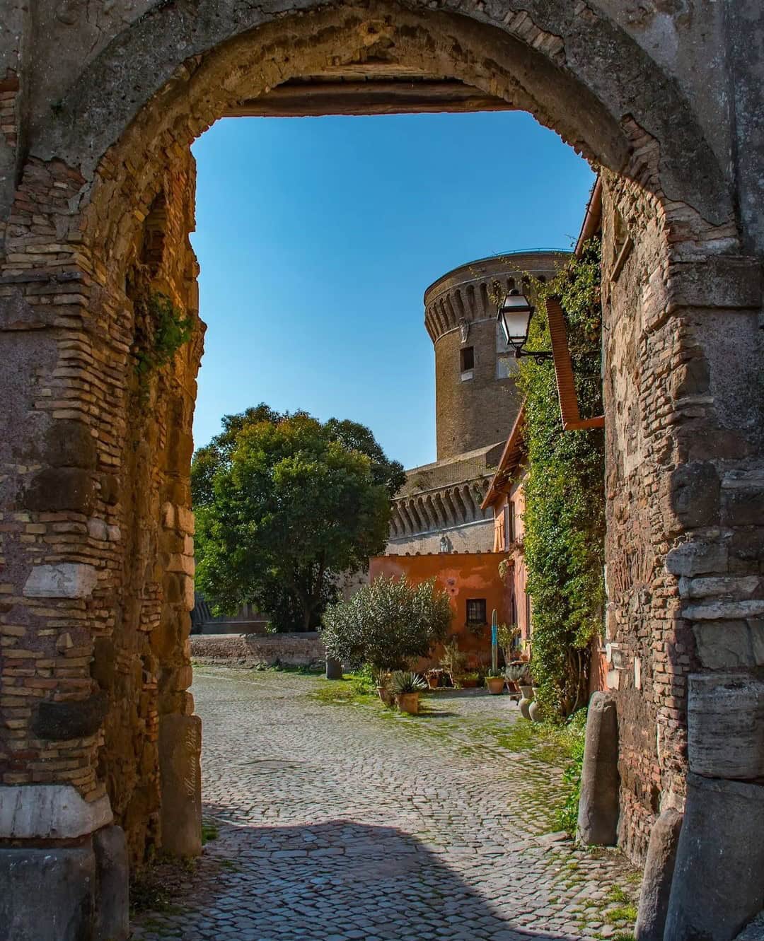 Stone archway opens to a cobblestone path, rustic houses, trees, and a round medieval tower under blue sky&mdash;one of the best day trips from Rome for families seeking charm and history.