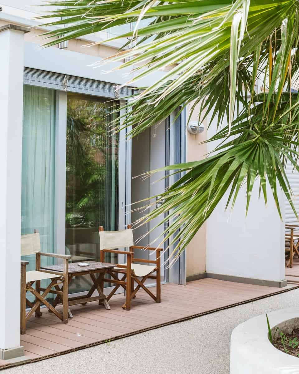 Two wooden chairs and a table sit on a patio outside a glass door, shaded by a large leafy palm tree&mdash;evoking the relaxed atmosphere of the best beach resorts in Italy for families.