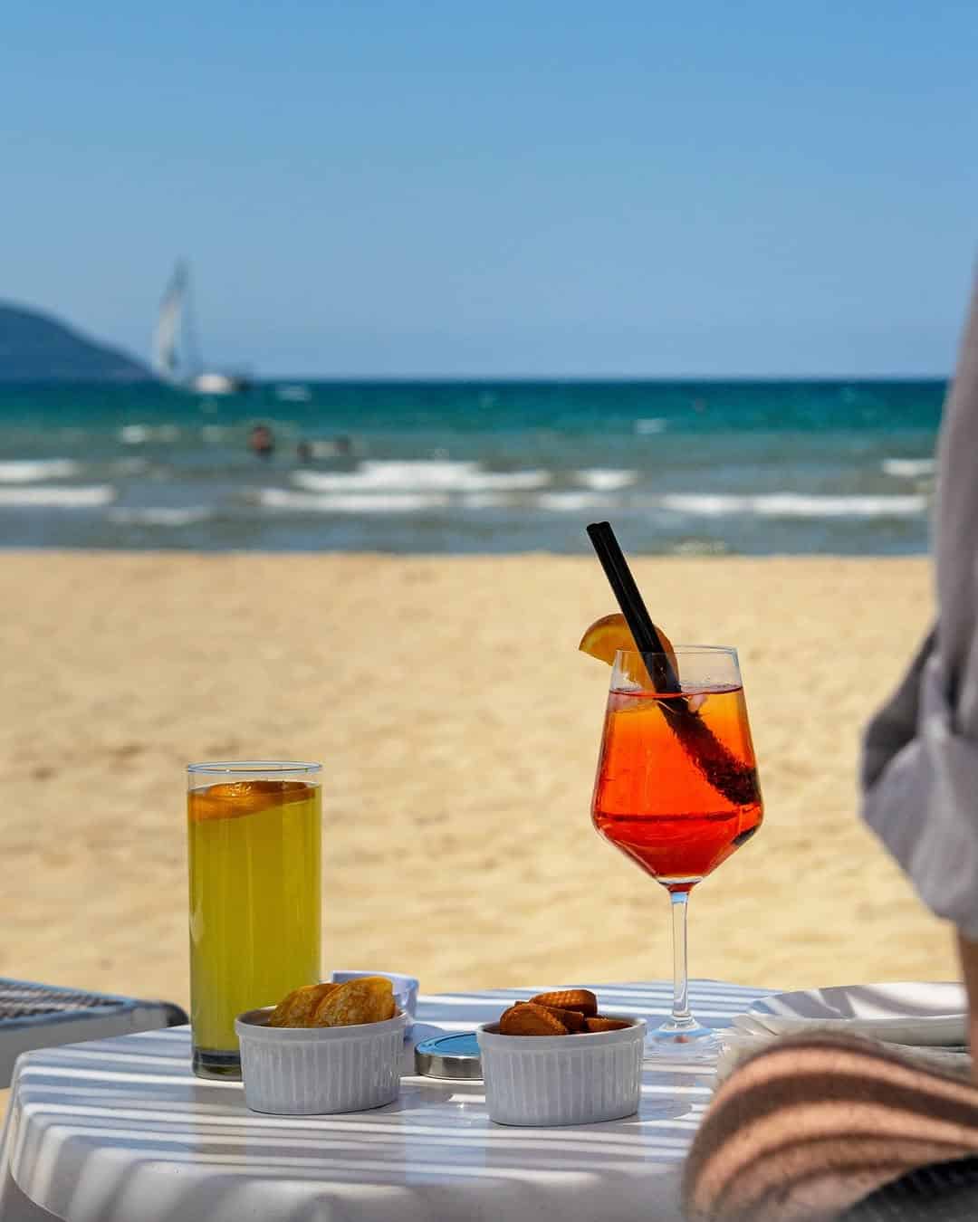 Two colorful drinks and snacks on a table by a sandy beach, with the sea and a sailboat in the background&mdash;perfect for relaxing moments at one of the best beach resorts in Italy for families.