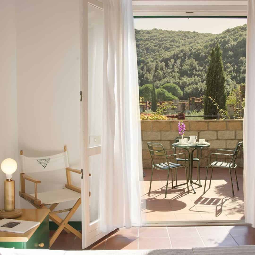 Sunlit terrace with green chairs and a table, reminiscent of the relaxed charm found at the best beach resorts in Italy for families, viewed from a cozy room with white curtains and a director&rsquo;s chair.