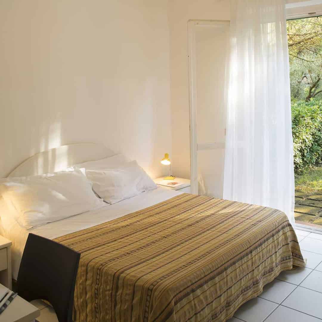 Bright bedroom with a double bed, striped blanket, and open door to a green garden&mdash;perfect for relaxing after exploring the best beach resorts in Italy for families.