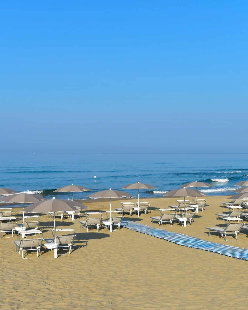 Rows of empty lounge chairs and umbrellas on a sandy beach at one of the best beach resorts in Italy for families, all facing a calm blue sea under a clear sky.