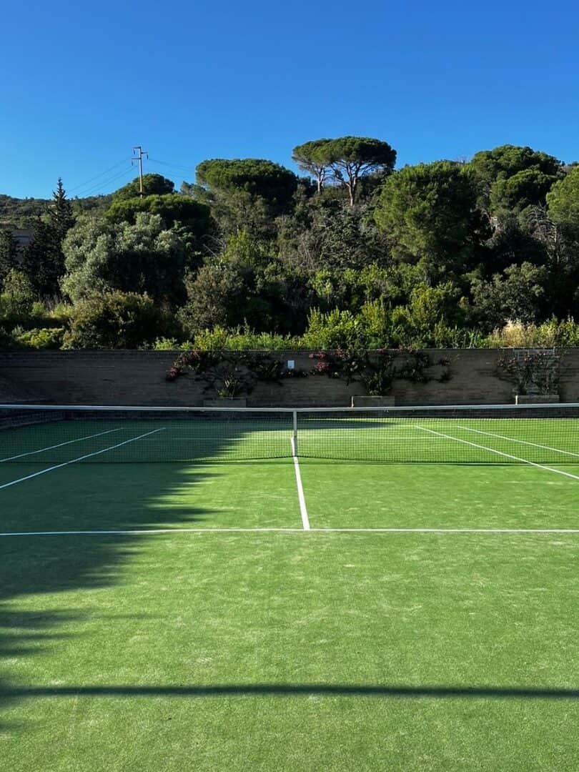 A green tennis court with white lines, surrounded by trees and hills under a clear blue sky&mdash;much like the views at the best beach resorts in Italy for families.