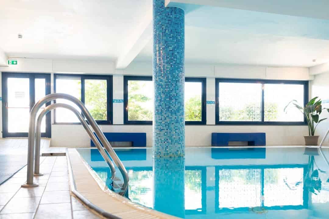 Indoor swimming pool with blue tile pillar, metal ladder, benches, and large windows letting in natural light&mdash;just like the relaxing amenities found at the best beach resorts in Italy for families.