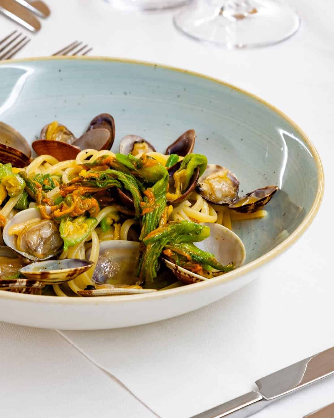 A bowl of seafood pasta with clams and zucchini flowers, served on a set table with cutlery and wine glasses&mdash;perfect for savoring the flavors found at the best beach resorts in Italy for families.