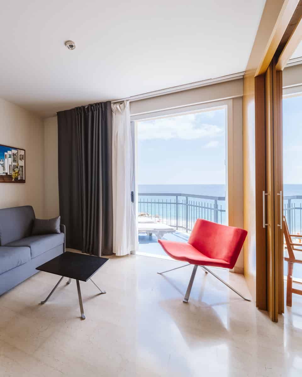 Modern living room with a gray sofa, red chair, black table, and balcony overlooking the ocean&mdash;perfect for relaxing after exploring the best beach resorts in Italy for families.