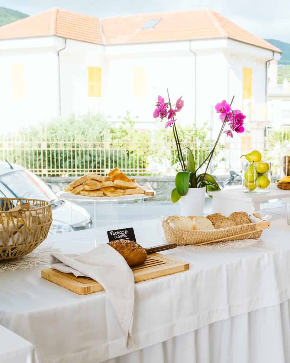 Breakfast buffet table with bread, pastries, fruit, and a pink orchid on a white tablecloth near a window&mdash;perfect for mornings at the best beach resorts in Italy for families.