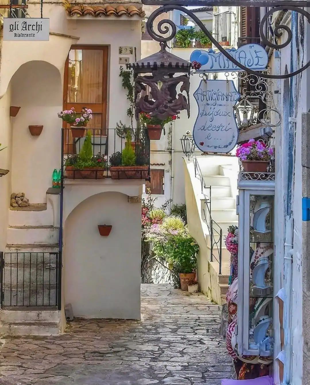Narrow cobblestone alley with potted flowers, white buildings, and a hanging restaurant sign in Italy&mdash;perfect for those seeking the best day trips from Rome for families.