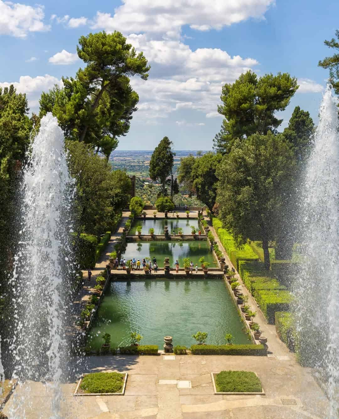 Large fountains frame a formal garden with rectangular ponds, lush trees, and people walking in the distance&mdash;one of the best day trips from Rome for families seeking beauty and relaxation.