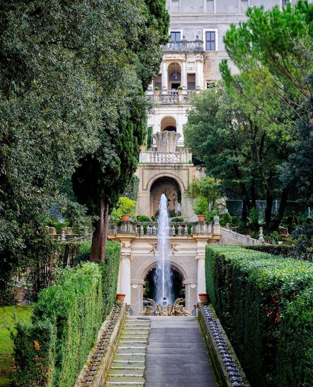 A tall fountain surrounded by trimmed hedges and trees, leading up to an ornate multi-story villa&mdash;a scene reminiscent of the best day trips from Rome for families seeking beauty and relaxation.