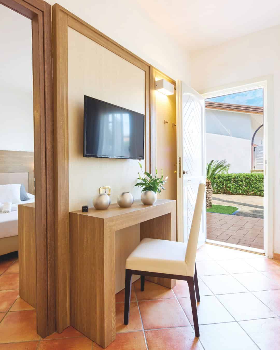 A small desk with a chair and TV in a bright room, reminiscent of the relaxing comforts found at the best beach resorts in Italy for families, with a door open to a sunny garden outside.