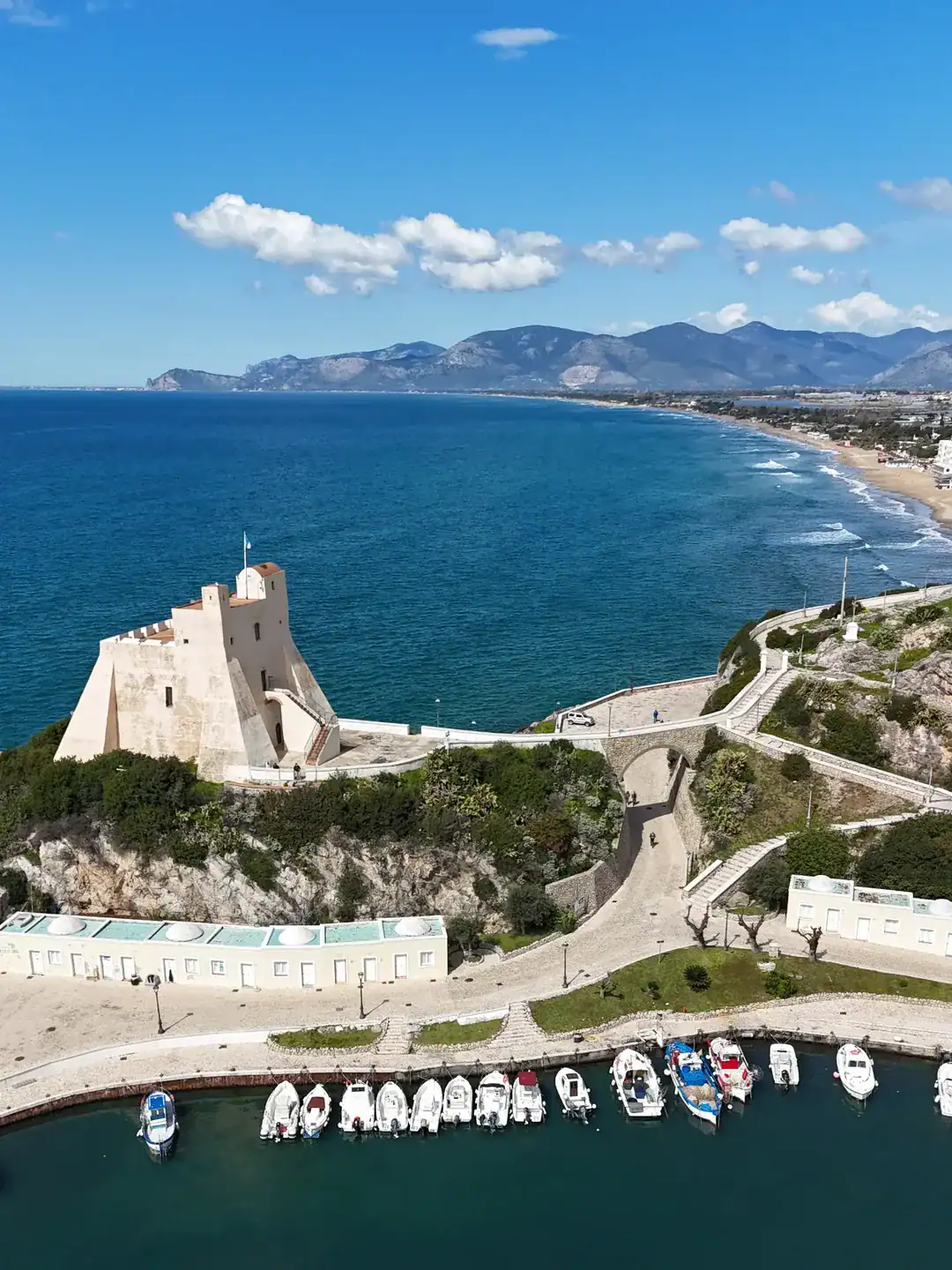 Aerial view of a seaside fortress, marina with boats, and coastline under a blue sky with scattered clouds&mdash;one of the best day trips from Rome for families.