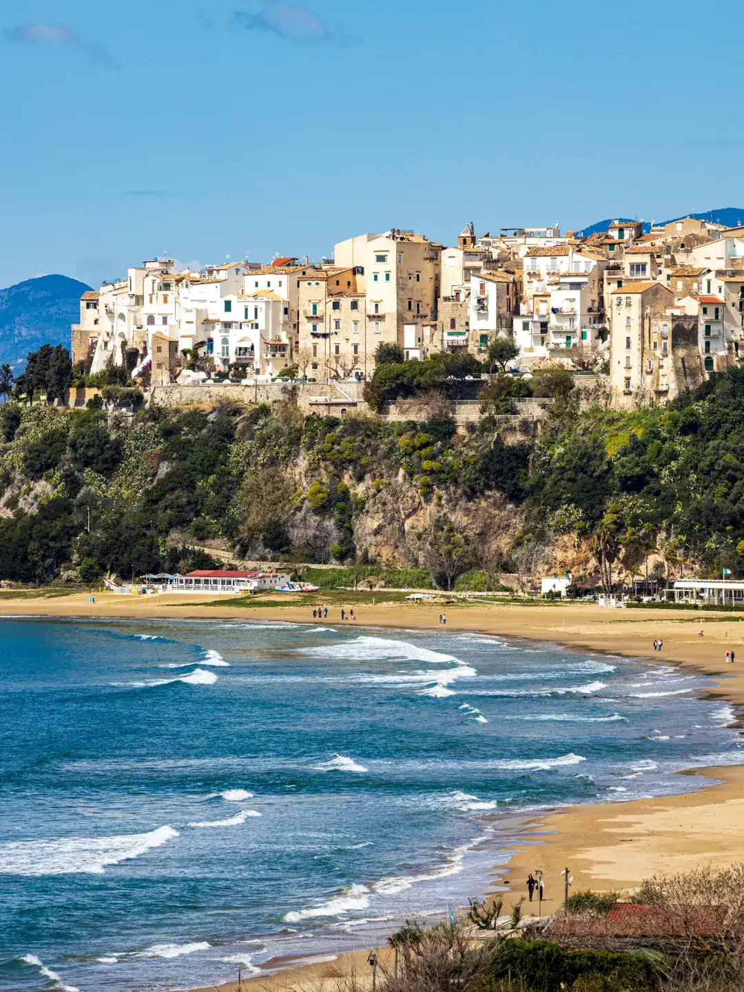Colorful cliffside buildings overlook a sandy beach with gentle waves and people walking along the shore, making it one of the best day trips from Rome for families.