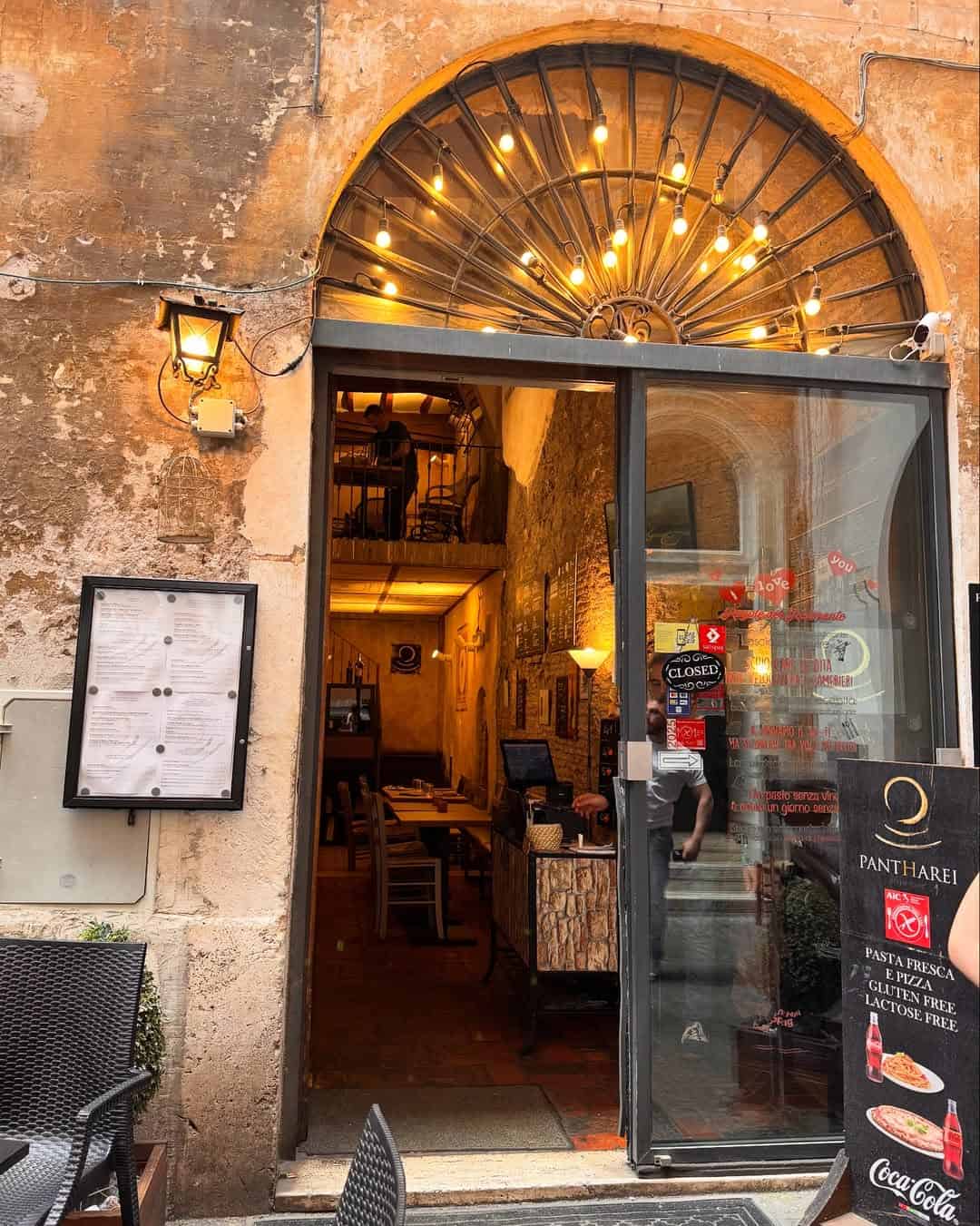 Cozy restaurant entrance with string lights, arched window, open door, and menu boards on a rustic stone wall—perfect for discovering some of the best Gluten Free Restaurants in Rome.