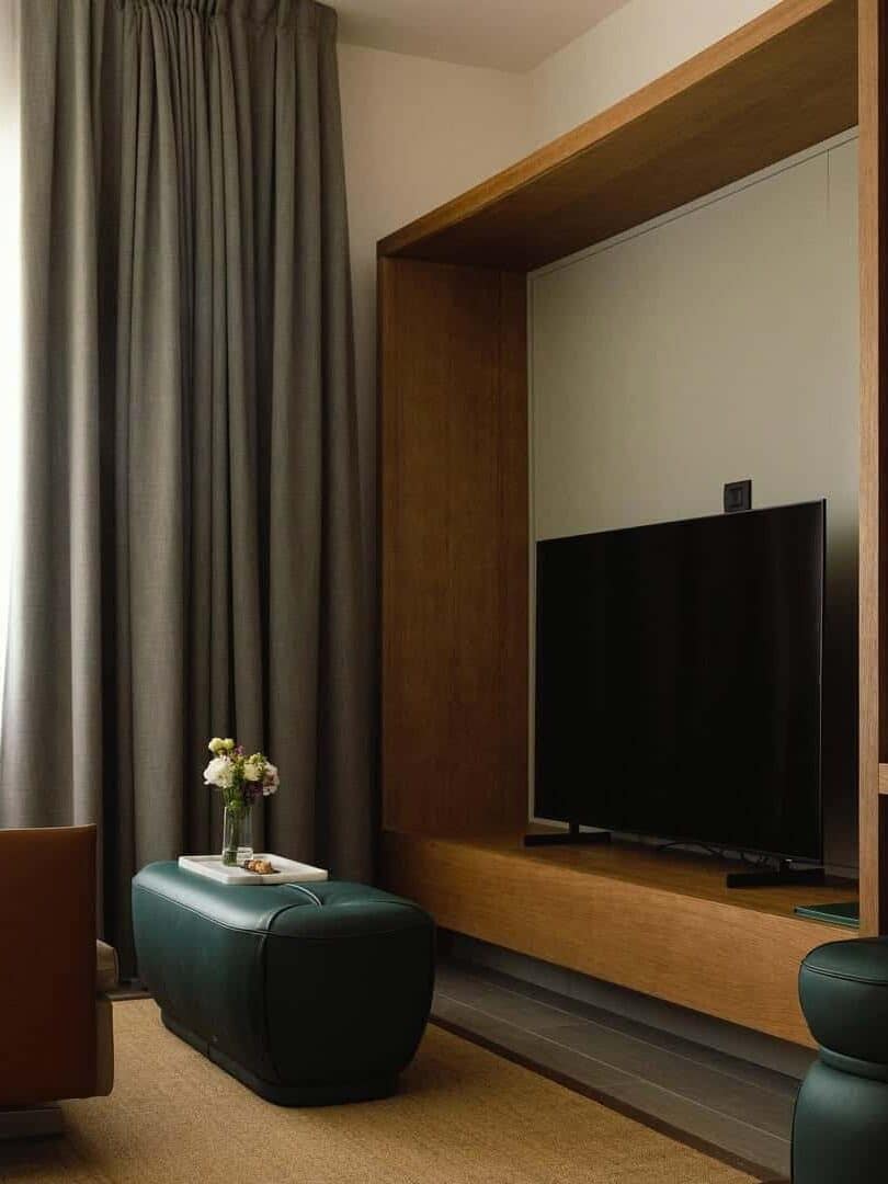 A modern living room with a TV, brown seating, green ottomans, and gray curtains by a large window&mdash;perfect for gathering family after sharing stories of the best beach resorts in Italy for families.