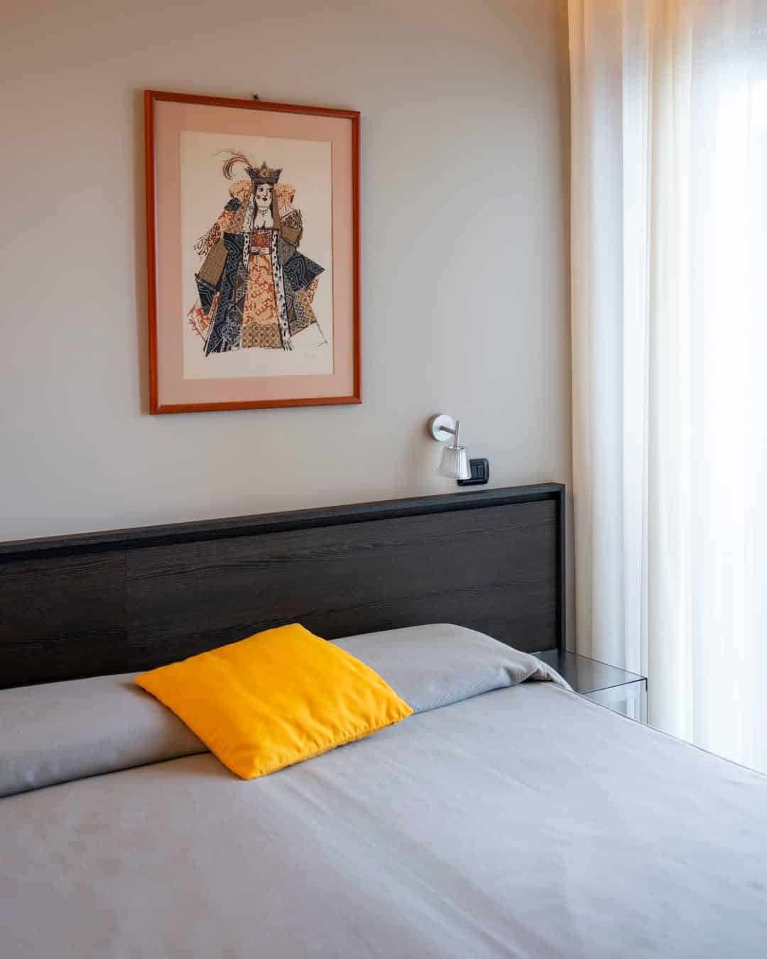 A neatly made bed with a yellow pillow, modern headboard, and framed art above it in a bright room, reminiscent of the inviting comfort found at the best beach resorts in Italy for families.