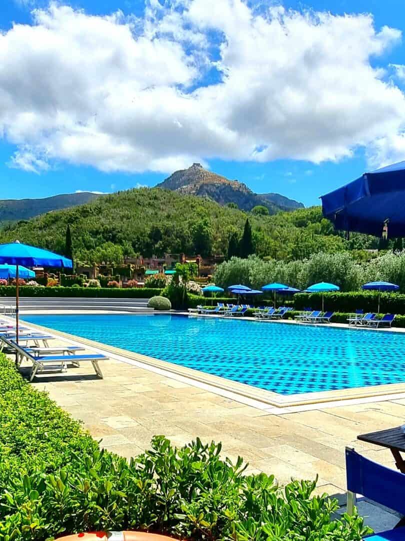 Outdoor swimming pool with blue umbrellas, lounge chairs, and mountains in the background under a partly cloudy sky&mdash;perfect for families seeking the best beach resorts in Italy for their next getaway.