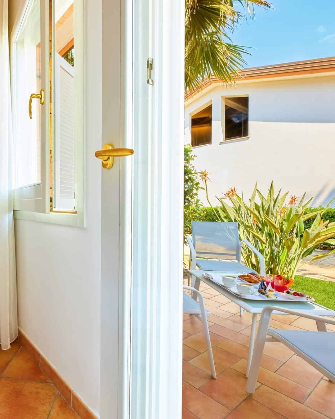 Open door to a sunny patio with two chairs, a small table with food, and lush green plants&mdash;reminiscent of relaxing mornings at the best beach resorts in Italy for families.