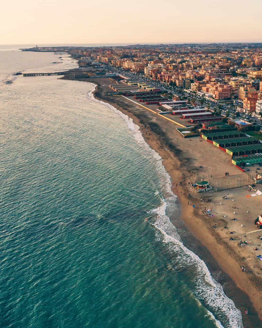 Aerial view of a city coastline with sandy beach, waves, and rows of buildings at sunset&mdash;an inviting destination reminiscent of the best day trips from Rome for families.