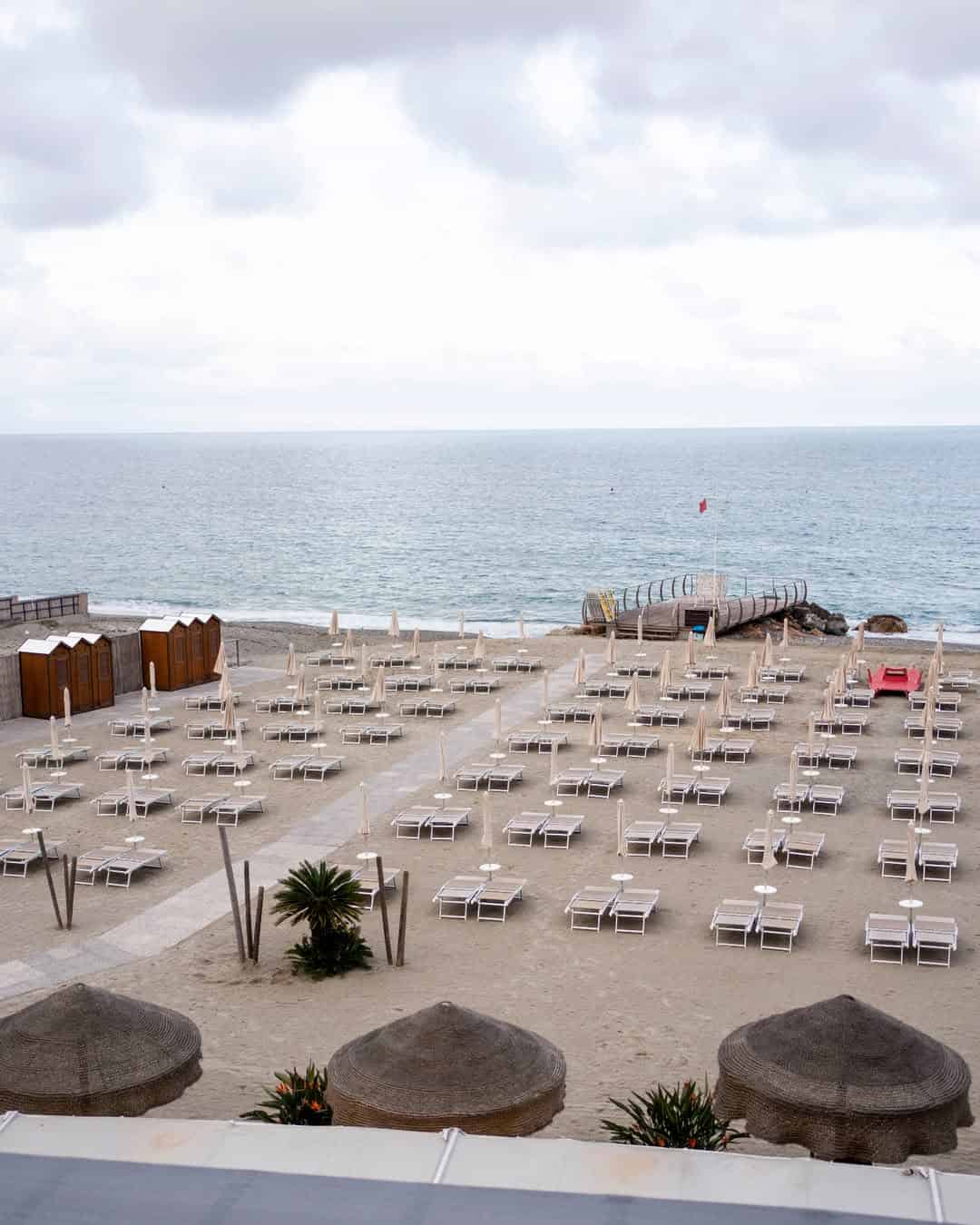 Rows of empty lounge chairs and umbrellas line the sandy beach by the sea under a cloudy sky, evoking the serene charm found at the best beach resorts in Italy for families.