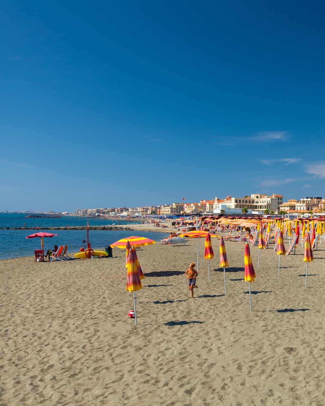 A sandy beach with colorful umbrellas, sun loungers, and families relaxing under a clear blue sky near a coastal town&mdash;one of the best day trips from Rome for families.