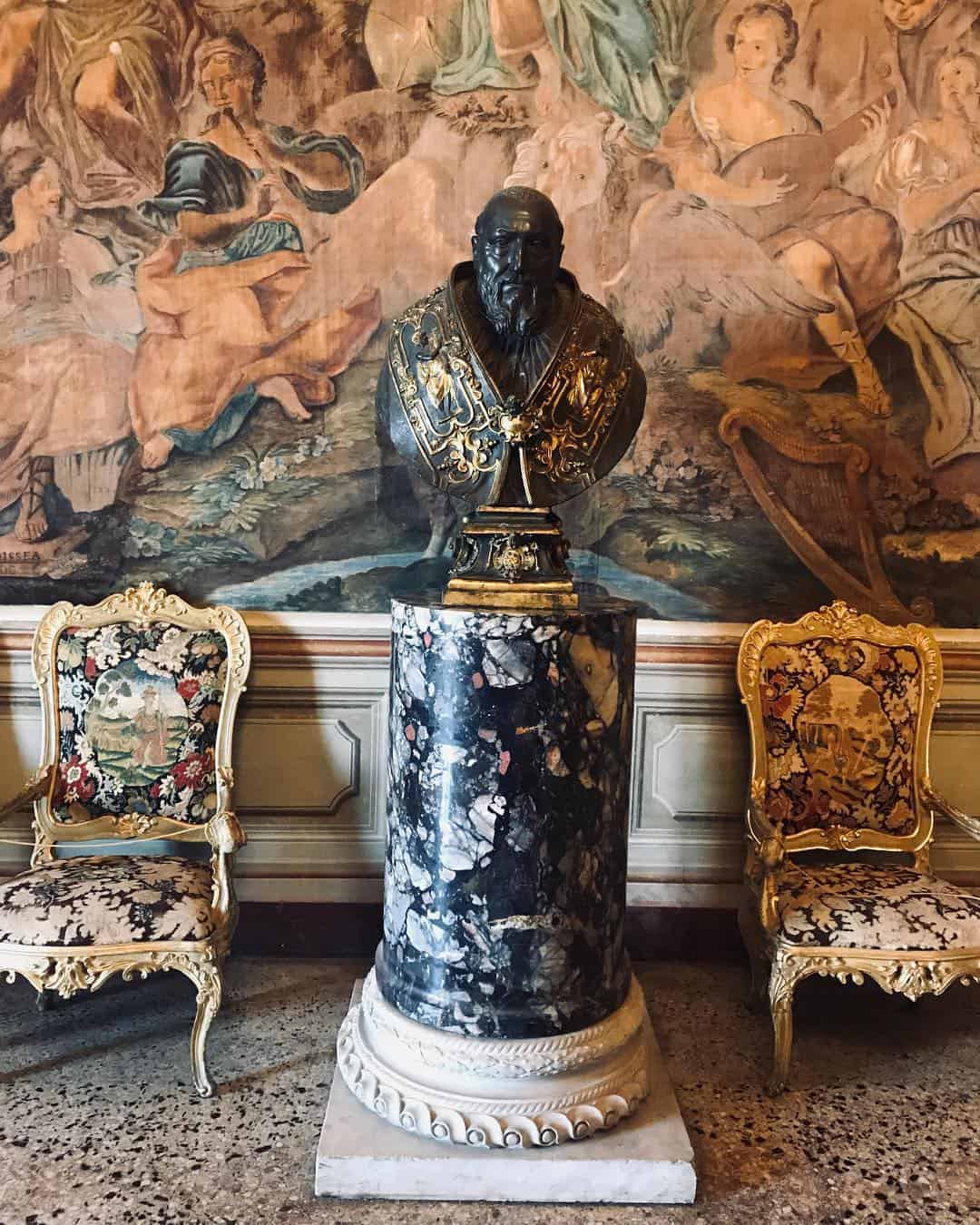 Marble bust on a pedestal between two ornate, floral-upholstered chairs with a detailed tapestry behind&mdash;an elegant scene reminiscent of the historic interiors you might discover on the best day trips from Rome for families.