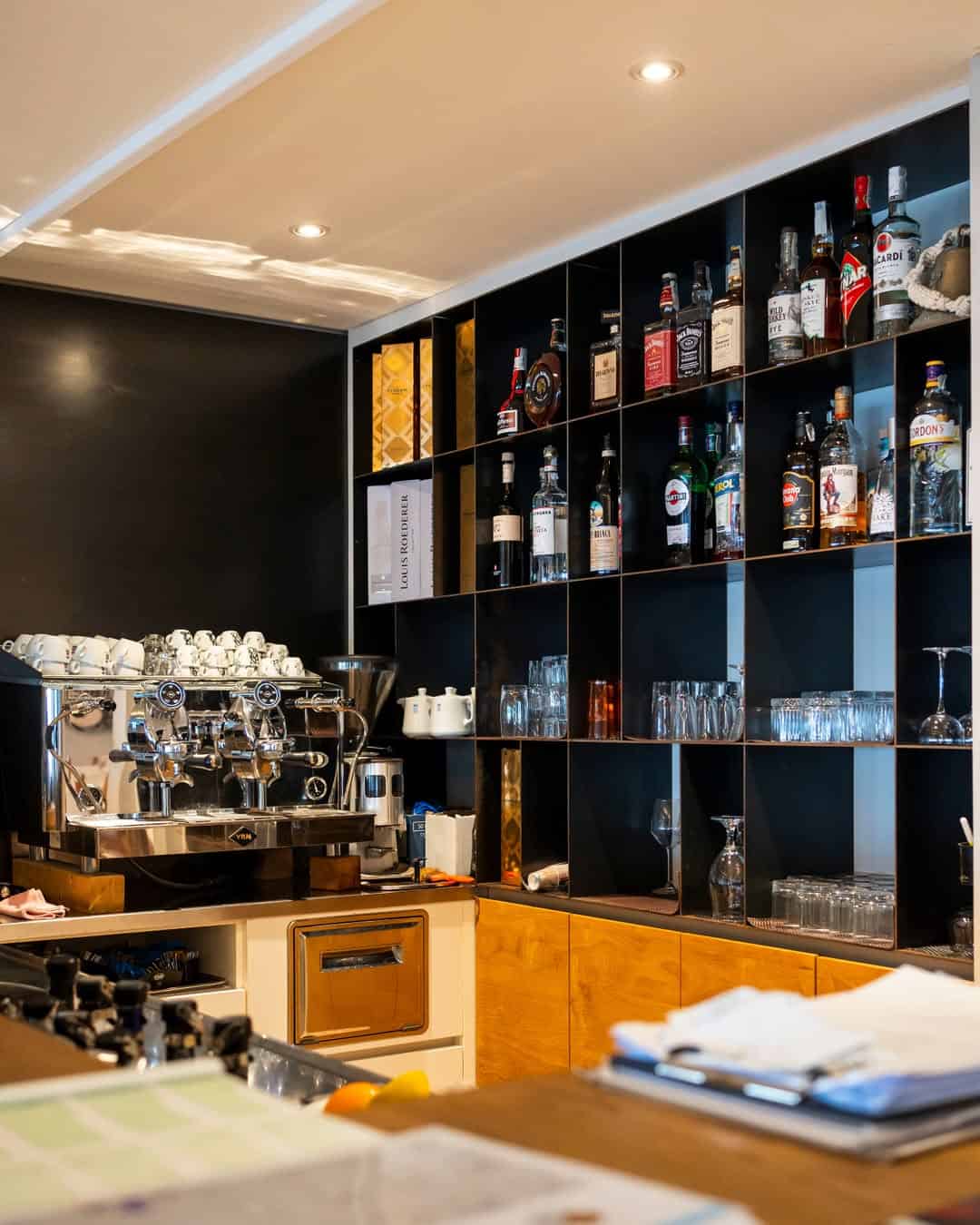 Modern bar with a coffee machine, glassware, and various liquor bottles organized on black shelves&mdash;much like the stylish lounges found at the best beach resorts in Italy for families.