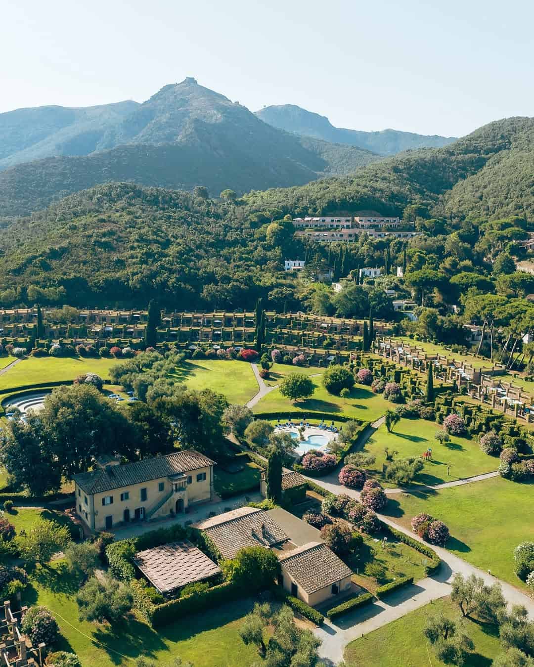 Aerial view of an estate with gardens, pools, and mountains in the background on a sunny day, reminiscent of the best beach resorts in Italy for families.