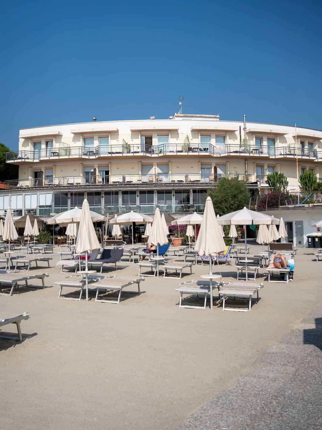 Sun loungers and umbrellas line the sandy beach in front of a modern, circular hotel&mdash;one of the best beach resorts in Italy for families&mdash;set beneath a clear blue sky.