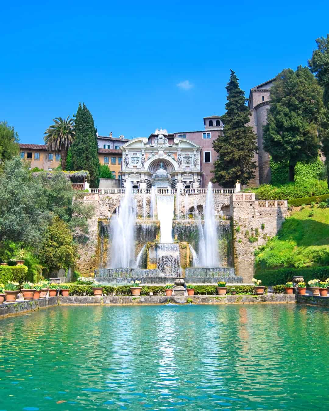 Elegant villa with cascading fountains and lush gardens reflected in a turquoise pool under a clear blue sky&mdash;one of the best day trips from Rome for families seeking relaxation and scenic beauty.