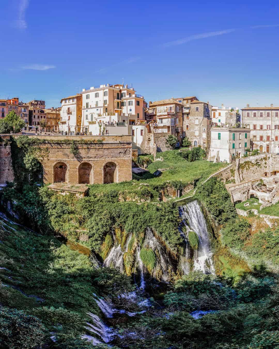 A small Italian town with historic buildings sits above a lush green waterfall and stone bridge under a blue sky, making it one of the best day trips from Rome for families seeking charm and natural beauty.