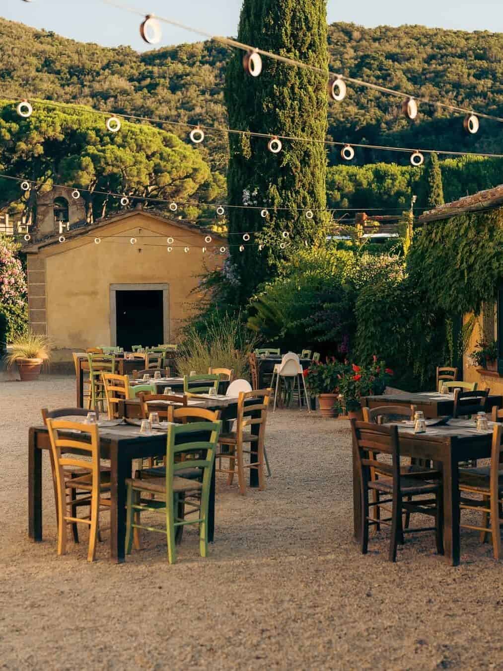 Outdoor cafe with wooden tables and chairs, string lights above, surrounded by greenery and hills&mdash;reminiscent of the relaxed charm found at the best beach resorts in Italy for families.