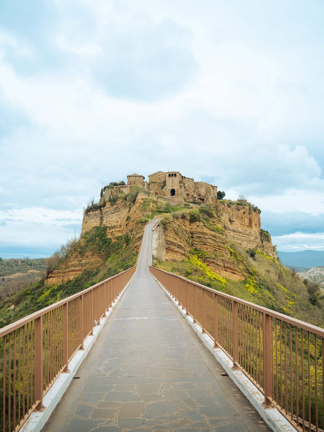 A long pedestrian bridge leads to a hilltop village with ancient stone buildings under a cloudy sky&mdash;one of the best day trips from Rome for families seeking history and stunning views.