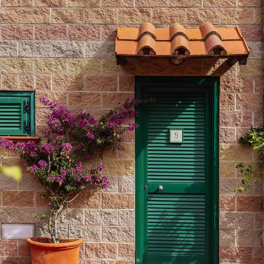 A green door with number 5, a potted flowering plant, and a small shuttered window on a brick wall evoke the charm found near the best beach resorts in Italy for families.