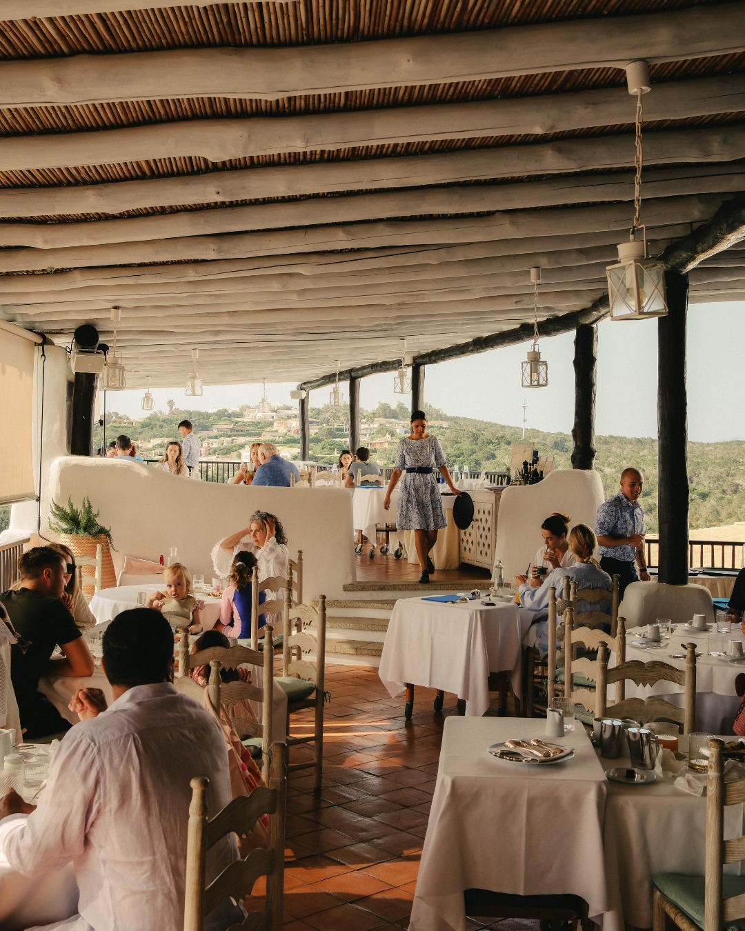 People dining at a rustic, open-air restaurant with a scenic view&mdash;perfect for those seeking the charm found at the best beach resorts in Italy for families&mdash;and a woman standing near a stage area.