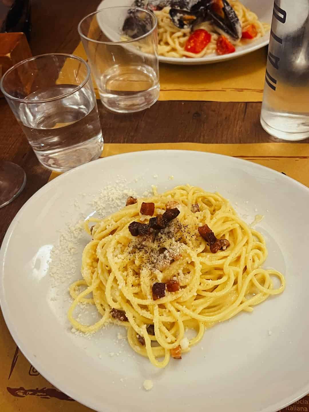 A plate of spaghetti carbonara with grated cheese and bacon, set on a table with water glasses and another pasta dish—perfect inspiration for those seeking Gluten Free Restaurants in Rome.
