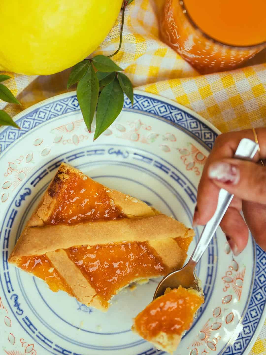 A hand holds a fork with a piece of apricot tart on a decorative plate beside a glass of orange juice—perfect inspiration for those seeking treats at Gluten Free Restaurants in Rome.