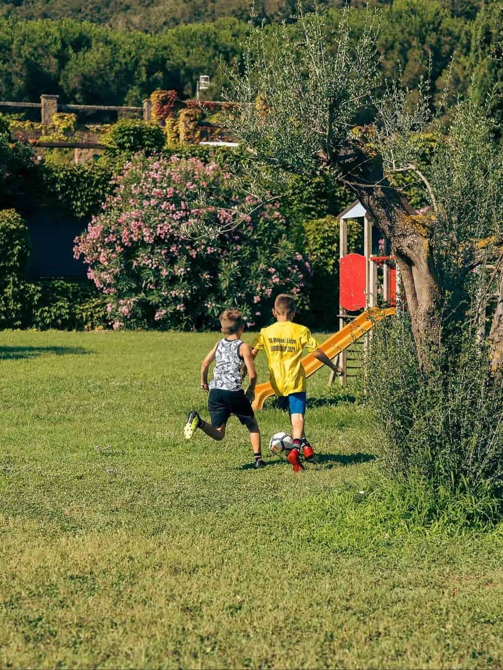 Two boys play soccer on a grassy field near a playground and trees on a sunny day, capturing the carefree spirit families enjoy at the best beach resorts in Italy for families.