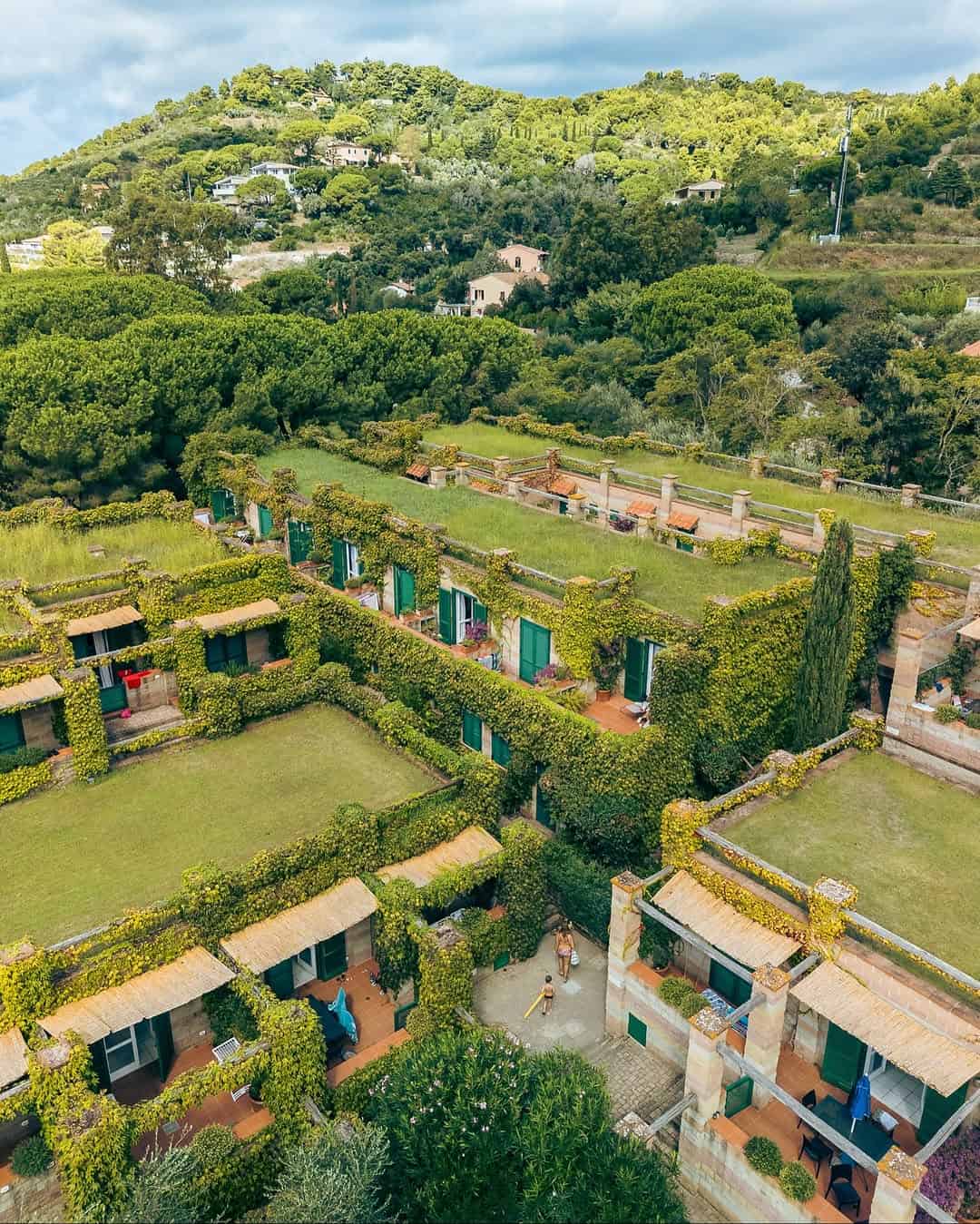 Aerial view of green-roofed buildings surrounded by lush trees and hills on a sunny day, reminiscent of the best beach resorts in Italy for families.