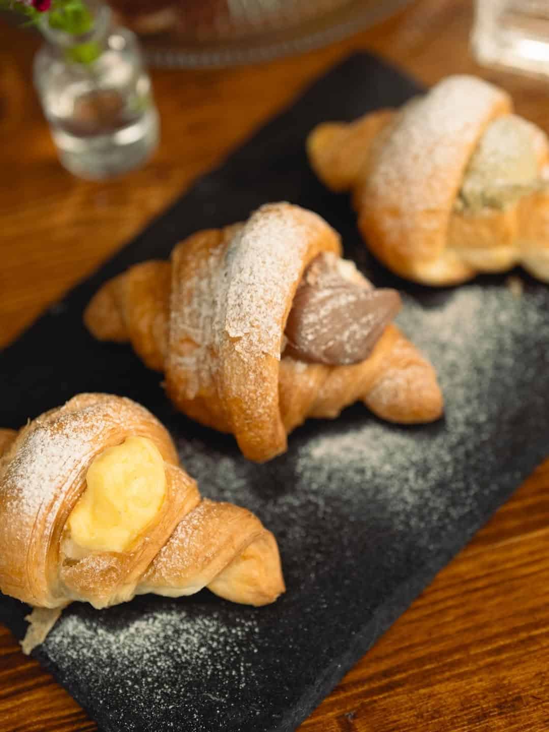 Three croissants filled with cream and chocolate, dusted with powdered sugar on a black slate platter—reminiscent of the pastries found in top gluten free restaurants in Rome.