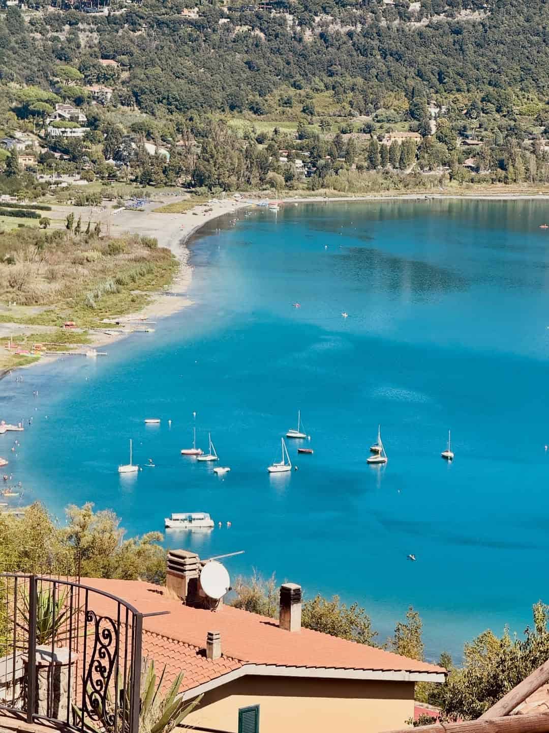 Turquoise lake with sailboats, surrounded by greenery and hills, seen from a balcony with terracotta rooftops&mdash;an idyllic view to enjoy during 4 days in Castel Gandolfo.