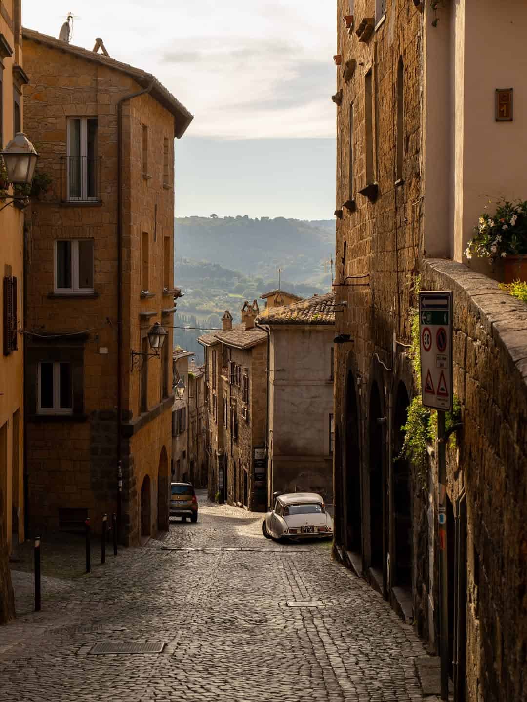 Narrow cobblestone street lined with old buildings and a parked car, set against hills in the distance&mdash;perfect for those seeking the best day trips from Rome for families.