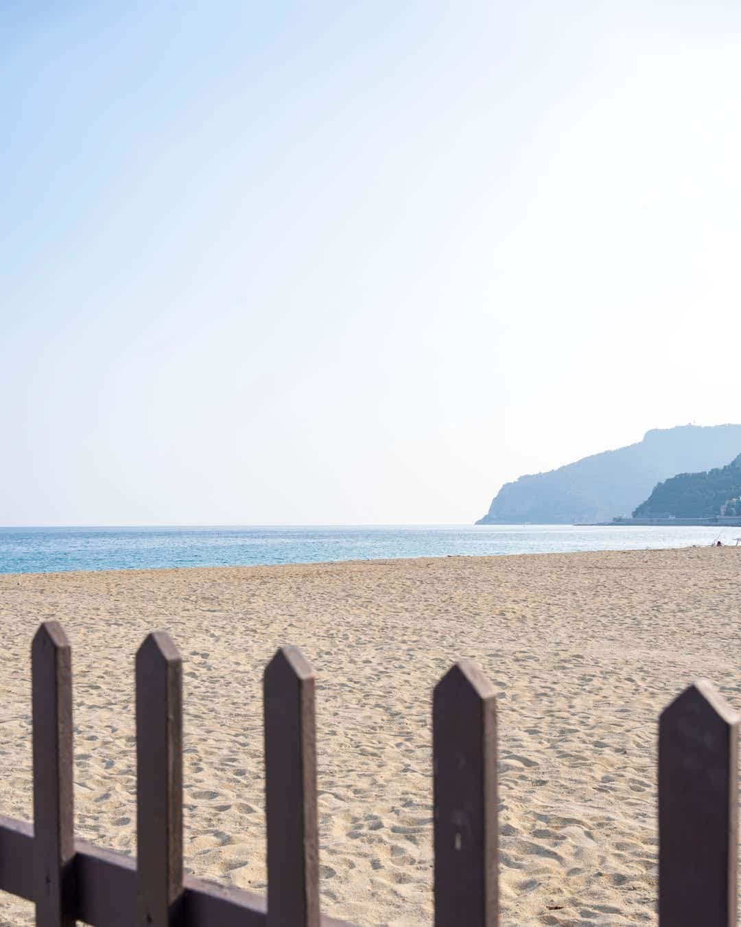 A sandy beach with a wooden fence in the foreground and distant hills by the sea under a clear sky&mdash;an inviting view reminiscent of the best beach resorts in Italy for families.
